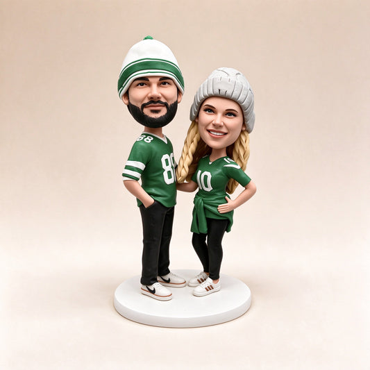 Two custom polymer clay figurines in green football jerseys and knit beanies standing on a white round base