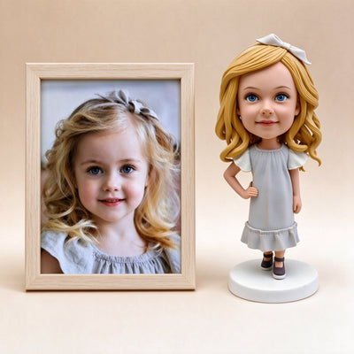 Framed photo of a young girl beside the custom polymer clay figurine made from it, in a light grey dress with a white bow